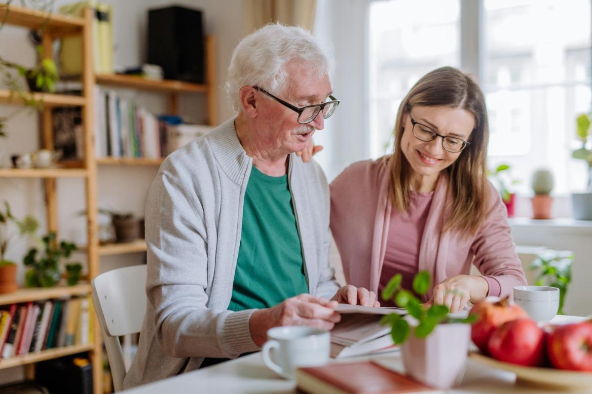 Elderly man and a caregiver reading a book together at a table.