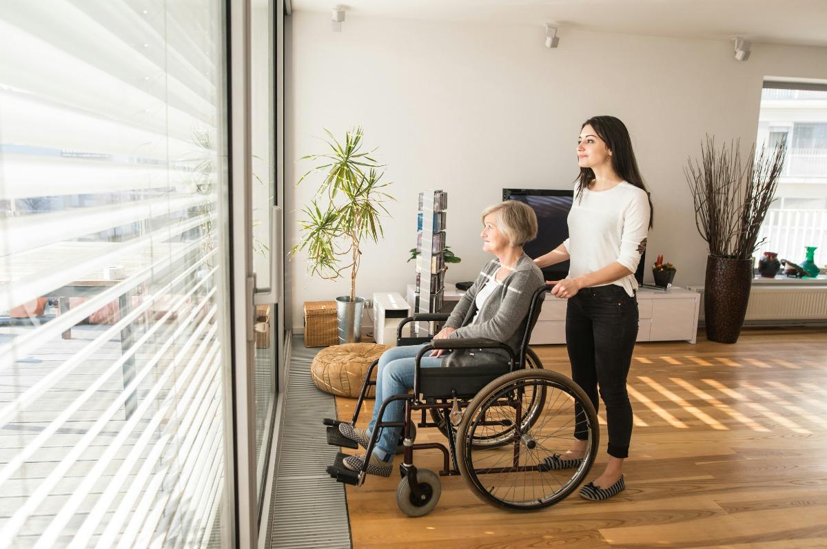 Caregiver pushing an elderly woman in a wheelchair near a window.