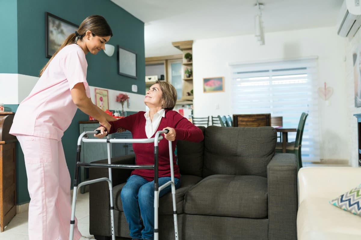 Caregiver helping a woman with a walker stand up from a chair.