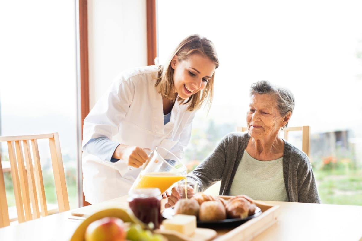 Caregiver pouring juice for an elderly woman at a dining table.