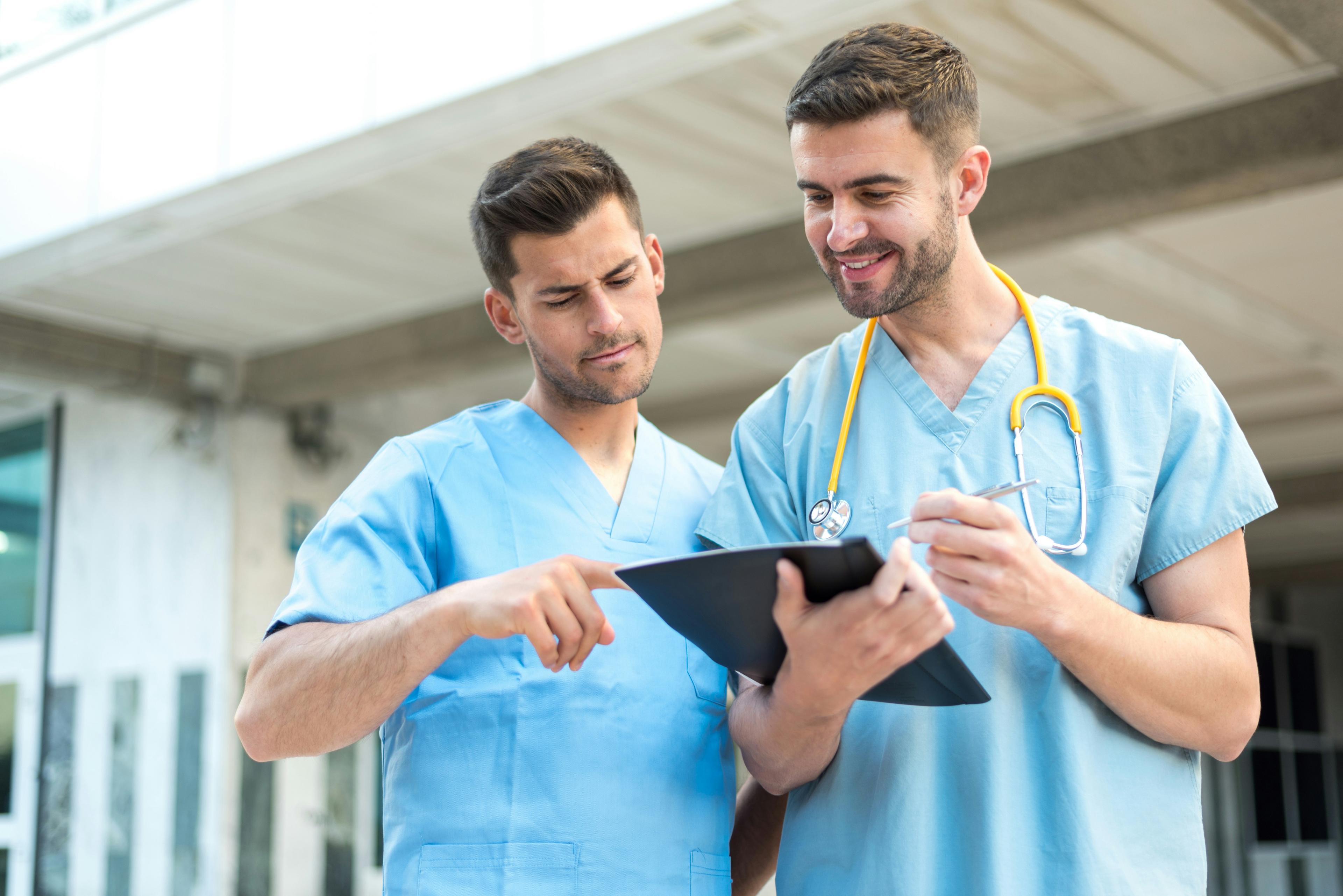 Two male nurses in blue scrubs discussing notes on a clipboard.