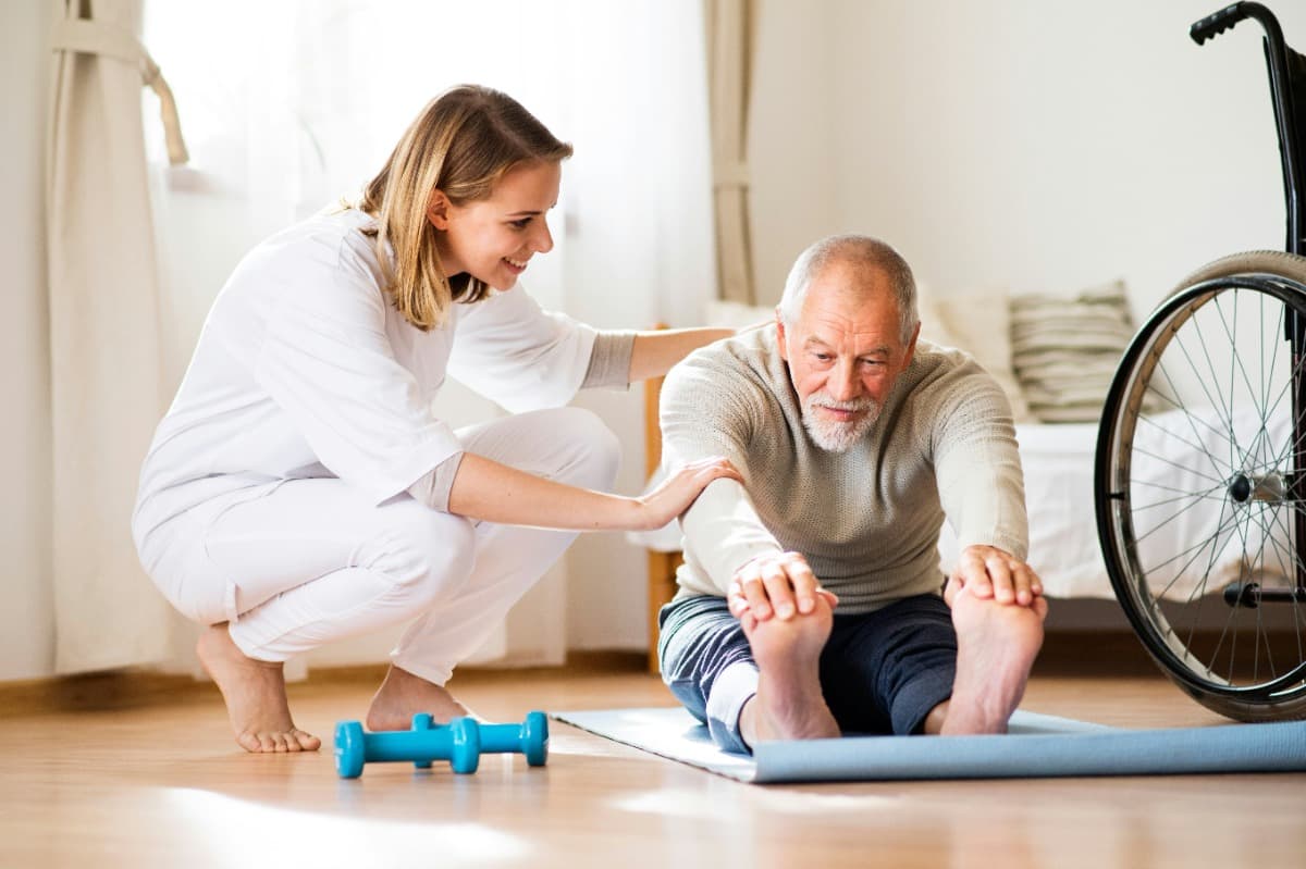 Caregiver assisting an elderly man with stretching exercises on a mat.