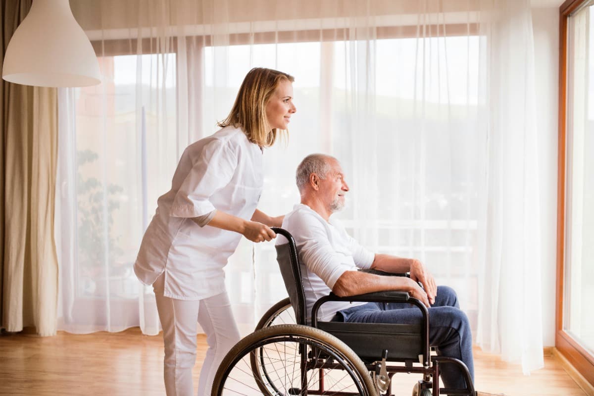 Caregiver pushing an elderly man in a wheelchair near a window.