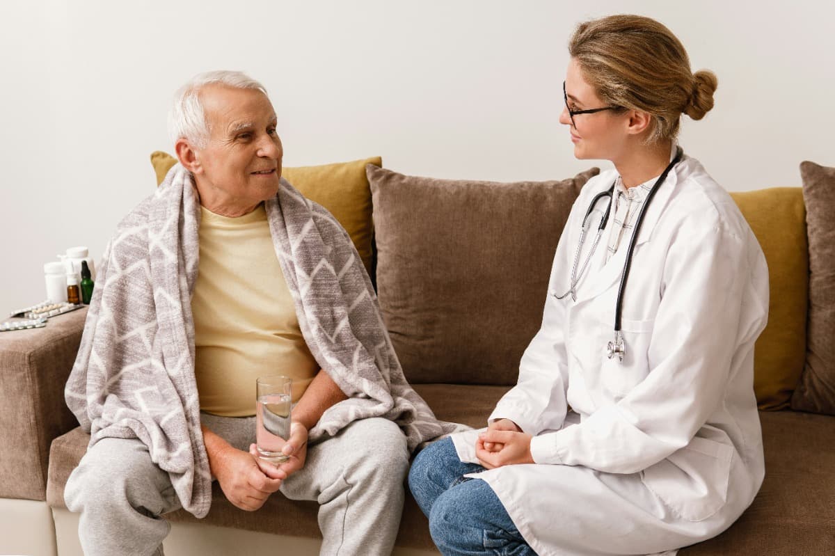 Elderly man sitting on a couch with a blanket talking to a nurse.