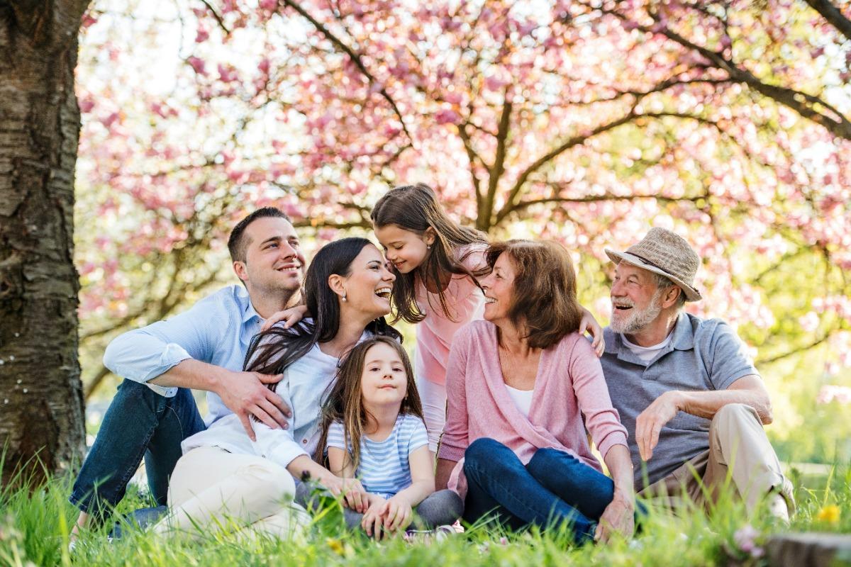 A happy multi-generational family sitting on grass in a park with blooming trees in the background.